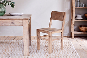 The Fioni Reclaimed Wood Dining Chair by nkuku sits by a matching wood table on a light patterned rug. A bowl and green vase with leaves rest atop, while shelves with bowls and plates complete the cozy farmhouse dining scene.