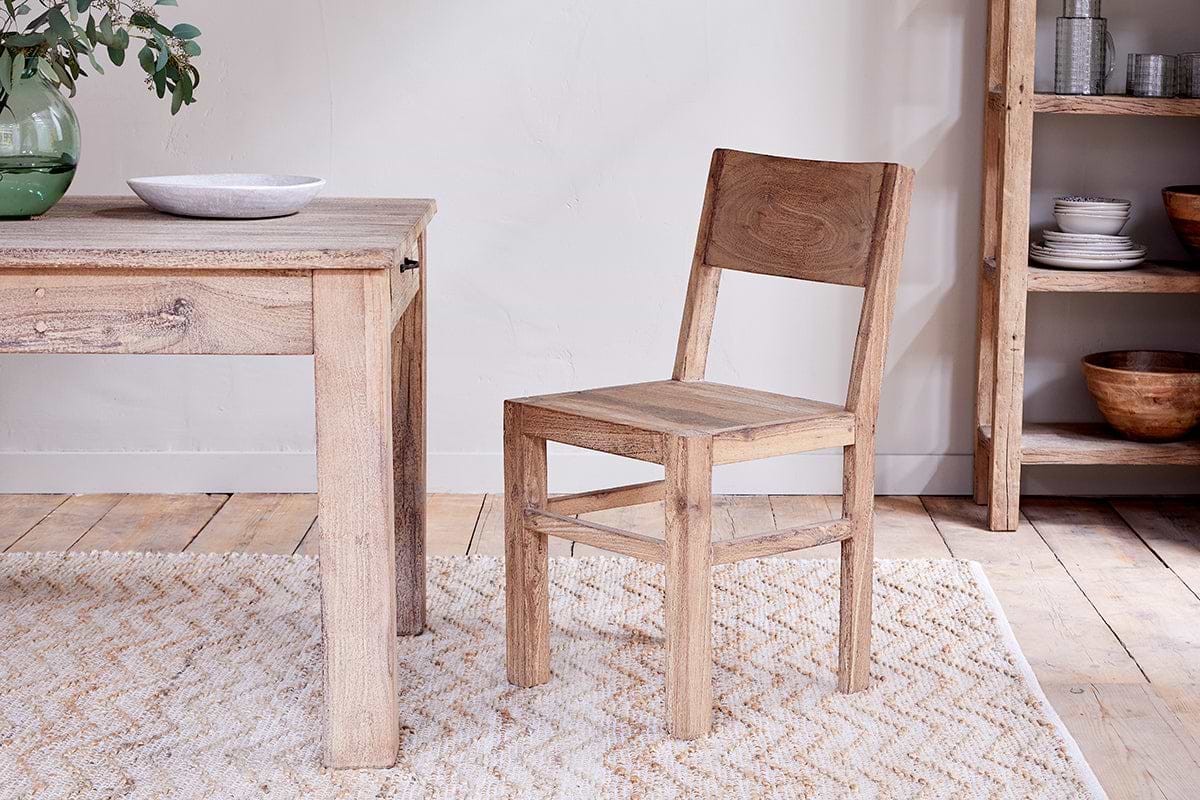 The Fioni Reclaimed Wood Dining Chair by nkuku sits by a matching wood table on a light patterned rug. A bowl and green vase with leaves rest atop, while shelves with bowls and plates complete the cozy farmhouse dining scene.