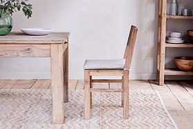 The Fioni Reclaimed Wood Dining Chair by nkuku, featuring a white cushion, pairs with a matching table on a light patterned rug. A shelf with dishes and a green vase brings freshness to this bright, minimalist farmhouse-inspired space.