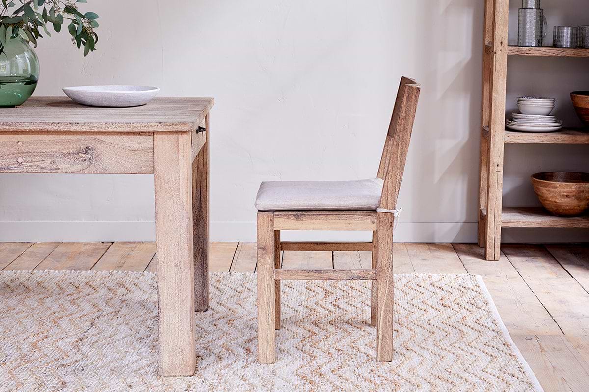 The Fioni Reclaimed Wood Dining Chair by nkuku, featuring a white cushion, pairs with a matching table on a light patterned rug. A shelf with dishes and a green vase brings freshness to this bright, minimalist farmhouse-inspired space.