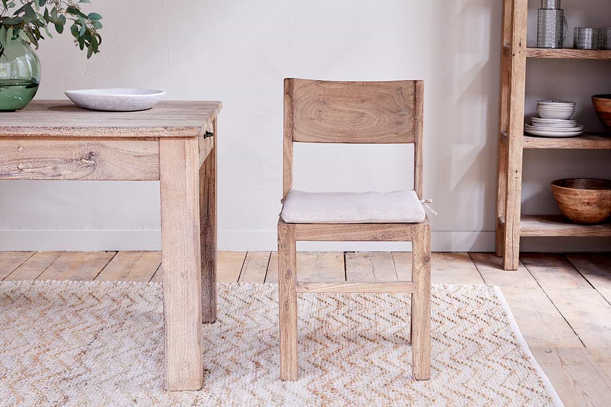 The nkuku Fioni Reclaimed Wood Dining Chair in Natural with a light cushion sits by a wooden table for a farmhouse look, while a woven rug lies beneath and a wooden shelf unit is visible on the right.