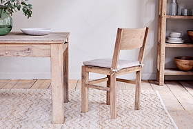 The nkuku Fioni Reclaimed Wood Dining Chair in natural, featuring a beige cushion, sits on a cream-patterned rug beside a matching wooden table. A wooden shelving unit with stacked bowls and plates and a green vase with branches complete the scene.