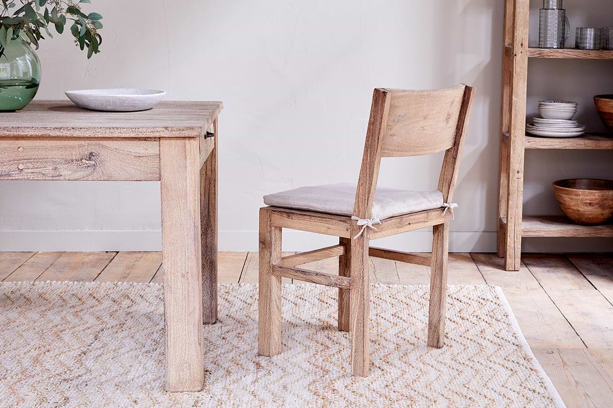 The nkuku Fioni Reclaimed Wood Dining Chair in natural, featuring a beige cushion, sits on a cream-patterned rug beside a matching wooden table. A wooden shelving unit with stacked bowls and plates and a green vase with branches complete the scene.