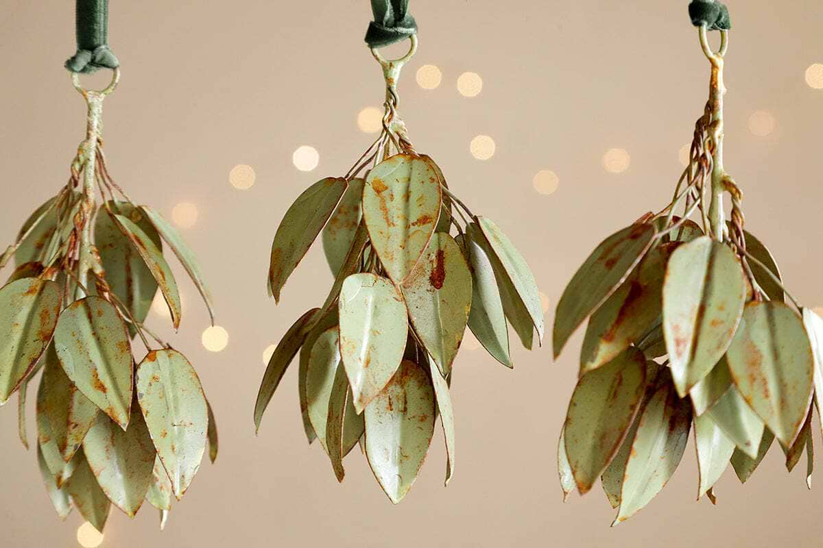 Four Geeti Baubles in soft green by nkuku hang from green velvet ribbons against a beige backdrop with blurred golden lights, adding a nature-inspired, serene touch with their elegant curves and subtle, organic hue.