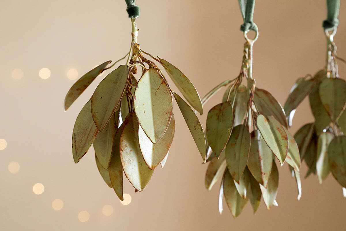 Four Geeti Baubles - Soft Green by nkuku hang from green ribbons against a soft beige background, with warm, blurred fairy lights. Their pale green finish and earthy tones evoke nature-inspired ornaments in this tranquil setting.