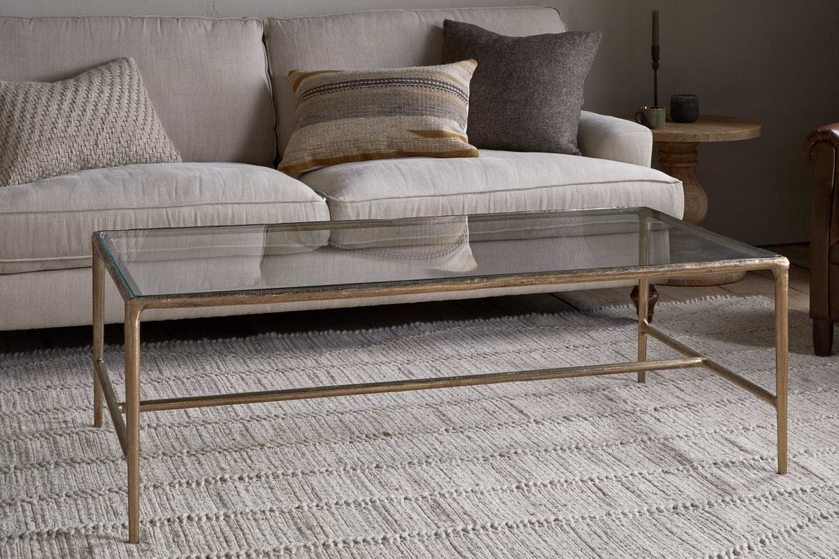 The nkuku Gilasi Cast Metal & Glass Coffee Table in antique brass sits on a textured cream rug before a light beige sofa with patterned cushions, creating a cozy living room setting.