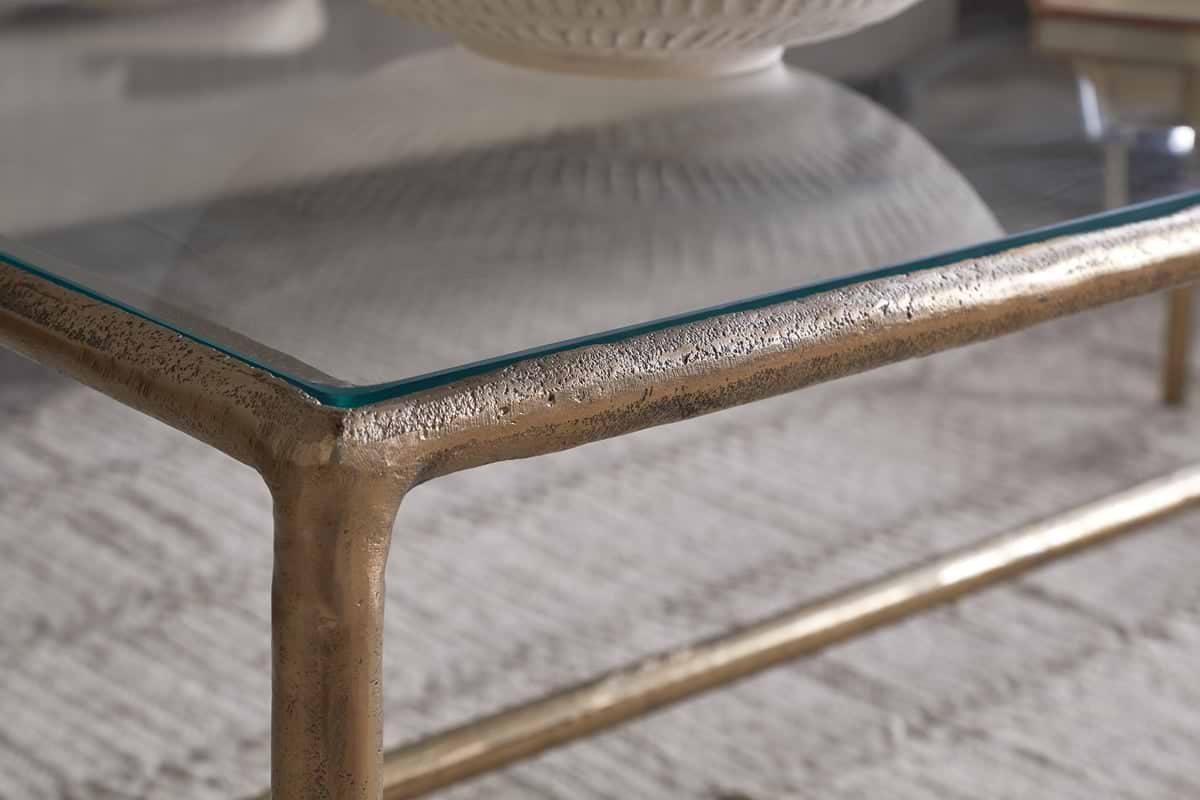 Close-up of the nkuku Gilasi Cast Metal & Glass Coffee Table in antique brass, featuring a textured metal frame and glass top on a light-patterned rug, with a white ceramic bowl partly visible on the tabletop.