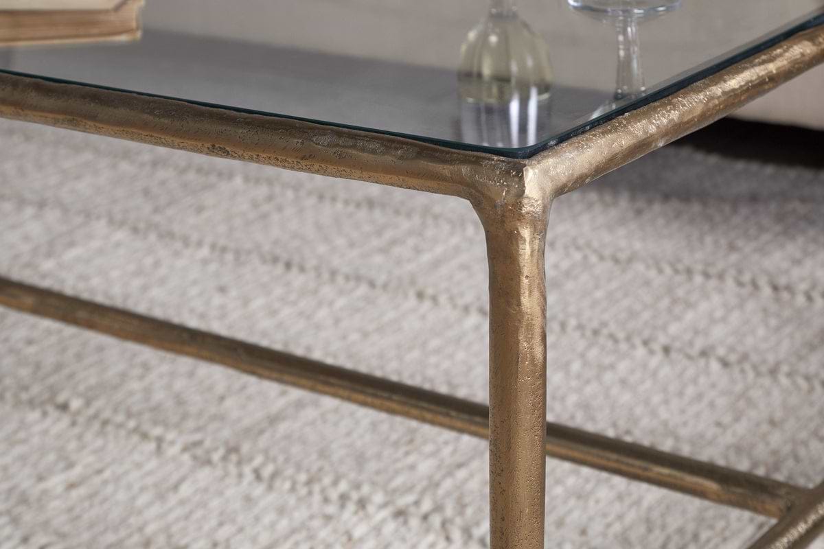 Close-up of the nkuku Gilasi Cast Metal & Glass Coffee Table in Antique Brass with a textured, hammered aluminium gold frame, set on a light-colored rug. A blurred glass bottle and glass appear in the background.