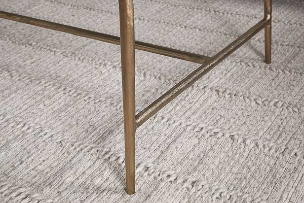 A close-up of the Gilasi Cast Metal & Glass Coffee Table by nkuku in antique brass, highlighting its leg with crossbar detail, standing on a textured, cream-colored woven rug with horizontal lines.