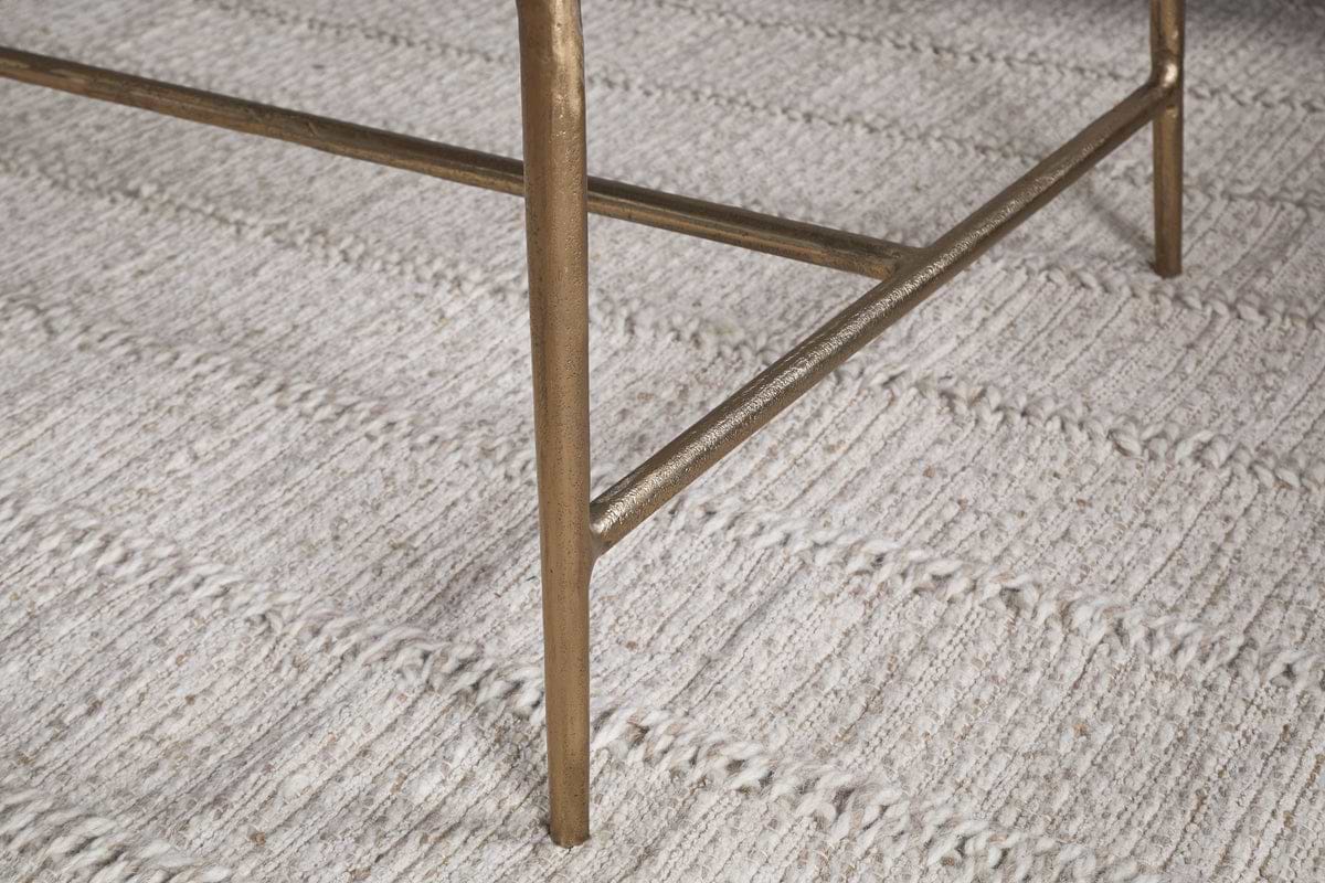 A close-up of the Gilasi Cast Metal & Glass Coffee Table by nkuku in antique brass, highlighting its leg with crossbar detail, standing on a textured, cream-colored woven rug with horizontal lines.