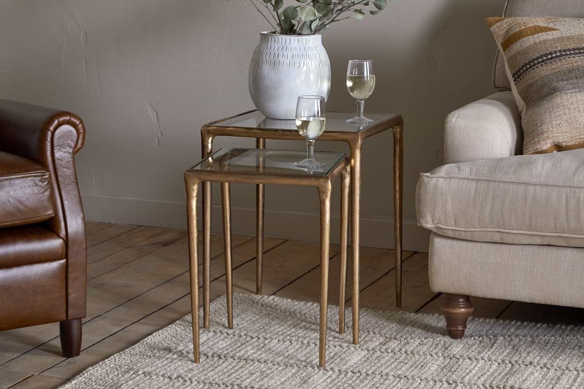 The nkuku Gilasi Cast Metal & Glass Nesting Side Tables in Antique Brass (Set of 2) display a vase with greenery and two white wine glasses, set between a brown leather chair and beige sofa on a light woven rug.