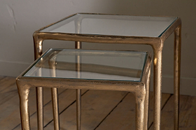 The nkuku Gilasi Cast Metal & Glass Nesting Side Tables (Set of 2) feature square glass tops and antique brass frames, with one table fitting neatly under the other, combining modern elegance and subtle vintage appeal.