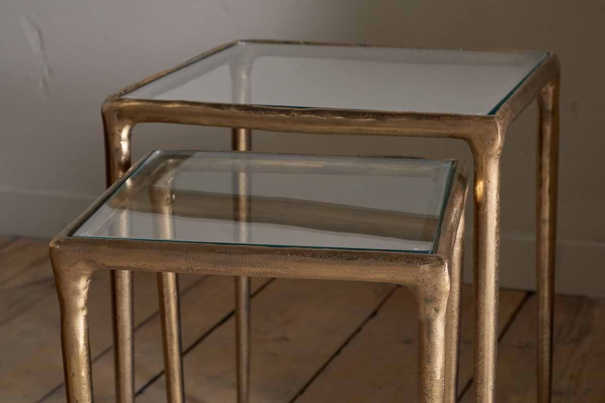 The nkuku Gilasi Cast Metal & Glass Nesting Side Tables (Set of 2) feature square glass tops and antique brass frames, with one table fitting neatly under the other, combining modern elegance and subtle vintage appeal.