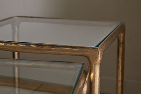 Close-up of nkukus Gilasi Cast Metal & Glass Nesting Side Tables (Set of 2) in antique brass, showcasing textured hammered aluminium frames, clear glass tops, and detailed corner and layered design against a neutral wall.