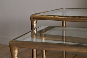 Close-up of the nkuku Gilasi Cast Metal & Glass Nesting Side Tables (Set of 2) in antique brass, featuring organic, irregular glass tops that evoke hammered aluminium. Shown on a wooden floor against a neutral wall.