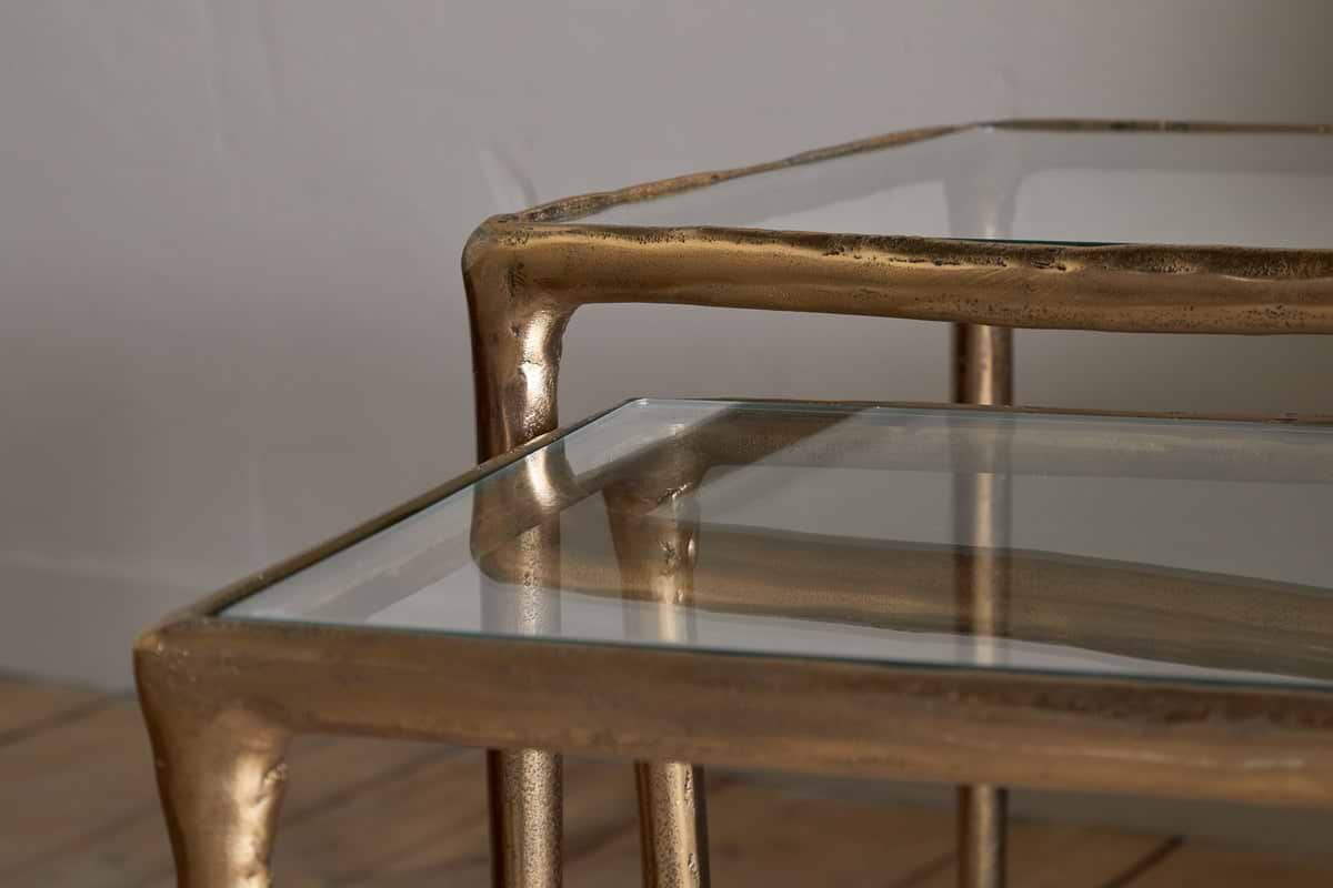 Close-up of the nkuku Gilasi Cast Metal & Glass Nesting Side Tables (Set of 2) in antique brass, featuring organic, irregular glass tops that evoke hammered aluminium. Shown on a wooden floor against a neutral wall.
