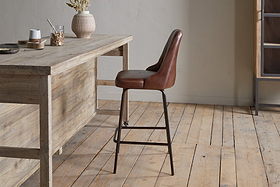 The nkuku Harsha Leather Counter Chair in chocolate brown, featuring black metal legs, provides stylish seating beside a rustic wooden table on a light wood floor in a minimalist room with neutral walls.