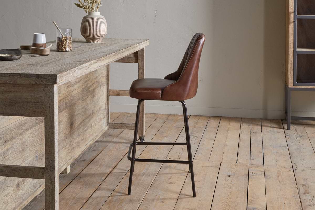 The nkuku Harsha Leather Counter Chair in chocolate brown, featuring black metal legs, provides stylish seating beside a rustic wooden table on a light wood floor in a minimalist room with neutral walls.