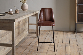 A modern kitchen features a wooden table on the left, ceramic vases and bowls, and the nkuku Harsha Leather Counter Chair in chocolate brown with black metal legs offering stylish seating at the island on a light wooden floor.