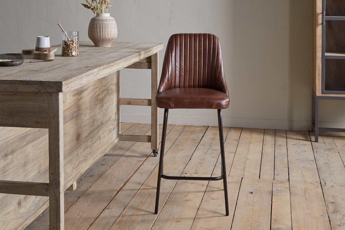 A modern kitchen features a wooden table on the left, ceramic vases and bowls, and the nkuku Harsha Leather Counter Chair in chocolate brown with black metal legs offering stylish seating at the island on a light wooden floor.