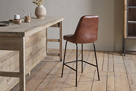 The Harsha Leather Counter Chair in chocolate brown by nkuku, featuring black metal legs, sits beside a rustic wooden table on light wood floors with neutral walls—ideal for stylish kitchen island seating. The table is adorned with a vase of dried flowers and jars.
