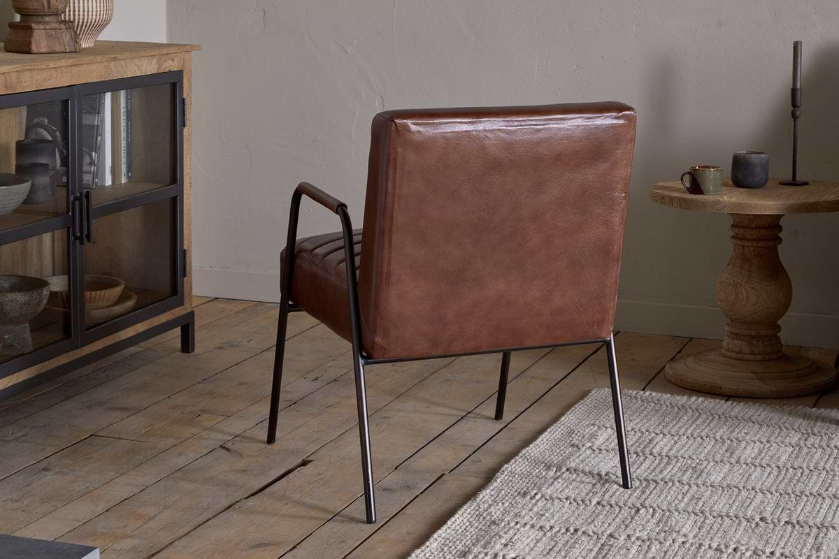 The nkuku Hadithi Leather Occasional Chair in chocolate brown, featuring a ribbed design and metal legs, sits on a light wooden floor near a beige textured rug alongside a wooden side table and glass-front cabinet against neutral walls.