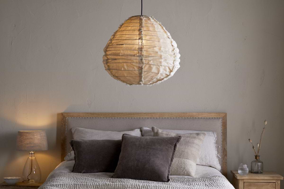 A cozy bedroom with a wooden bed frame, neutral bedding, dark throw pillows, and a nkuku Hirana Linen Lampshade in Natural (Small) hanging above. A nightstand with a small bedroom light and decor sits nearby.