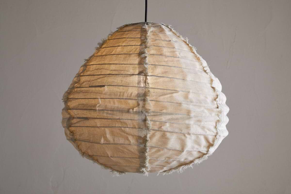 The Hirana Linen Lampshade by nkuku in natural, small size hangs from the ceiling, its textured shade softly glowing against a plain light gray background.