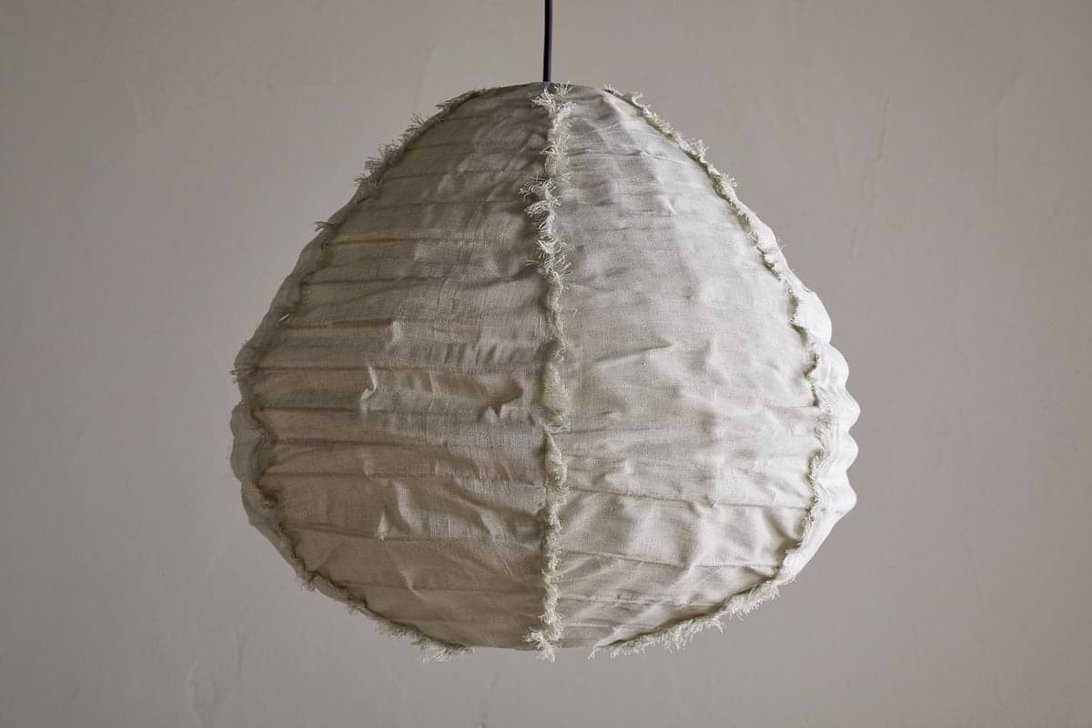 The Hirana Linen Lampshade - Natural - Small by nkuku, shaped like a teardrop with textured off-white linen, visible seams, and frayed edges, hangs from a slim black cord—an ideal bedroom pendant light against a plain wall.