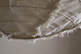 Close-up of the edge of the Hirana Linen Lampshade - Natural - Small by nkuku, showing its beige, textured linen fabric with visible fibers and loose threads against a soft, neutral background.