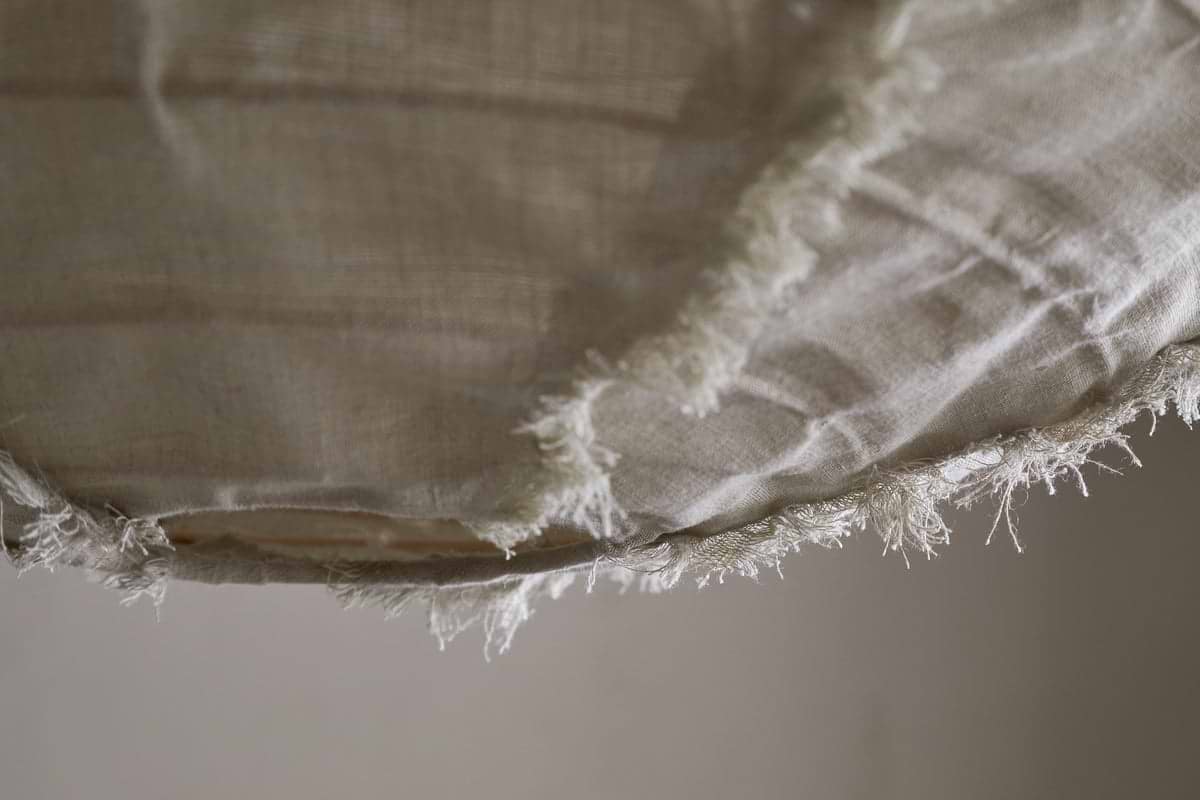 Close-up of the edge of the Hirana Linen Lampshade - Natural - Small by nkuku, showing its beige, textured linen fabric with visible fibers and loose threads against a soft, neutral background.
