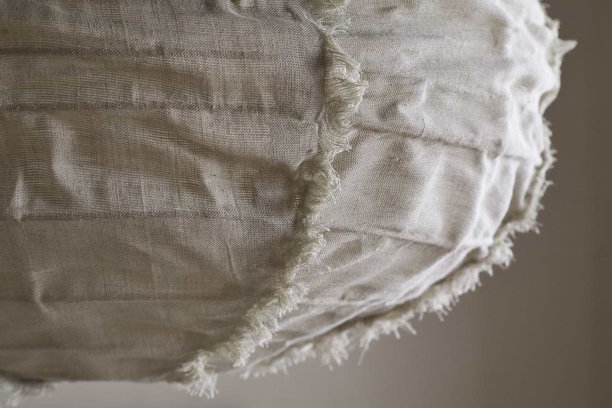 Close-up of the nkuku Hirana Linen Lampshade - Natural - Small, with visible seams and frayed edges. Its off-white textured linen and stitching create a rustic, handmade look—ideal for bedroom or pendant lighting. Background is softly blurred.