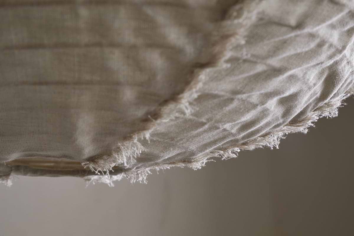 Close-up of the Hirana Cotton Lampshade by nkuku in natural, large size: off-white textured cotton with frayed edges and visible stitching, softly draped—perfect for adding warmth to any bedroom or living space.