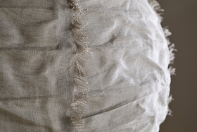 Close-up of the Hirana Cotton Lampshade by nkuku in Natural (Large), showing its light beige textured cotton fabric with a vertical seam and frayed edges, softly illuminated to highlight its unique details.