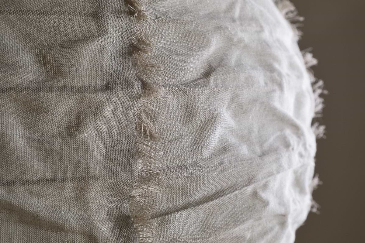 Close-up of the Hirana Cotton Lampshade by nkuku in Natural (Large), showing its light beige textured cotton fabric with a vertical seam and frayed edges, softly illuminated to highlight its unique details.