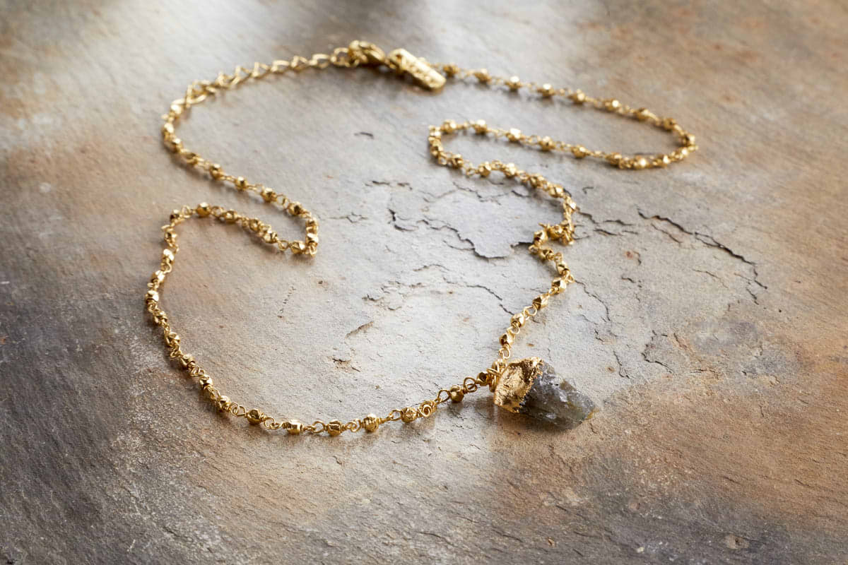 The nkuku Hadari Labradorite Necklace - Gold features a rough gray labradorite stone and delicate bead details, displayed on a textured rustic brown surface.