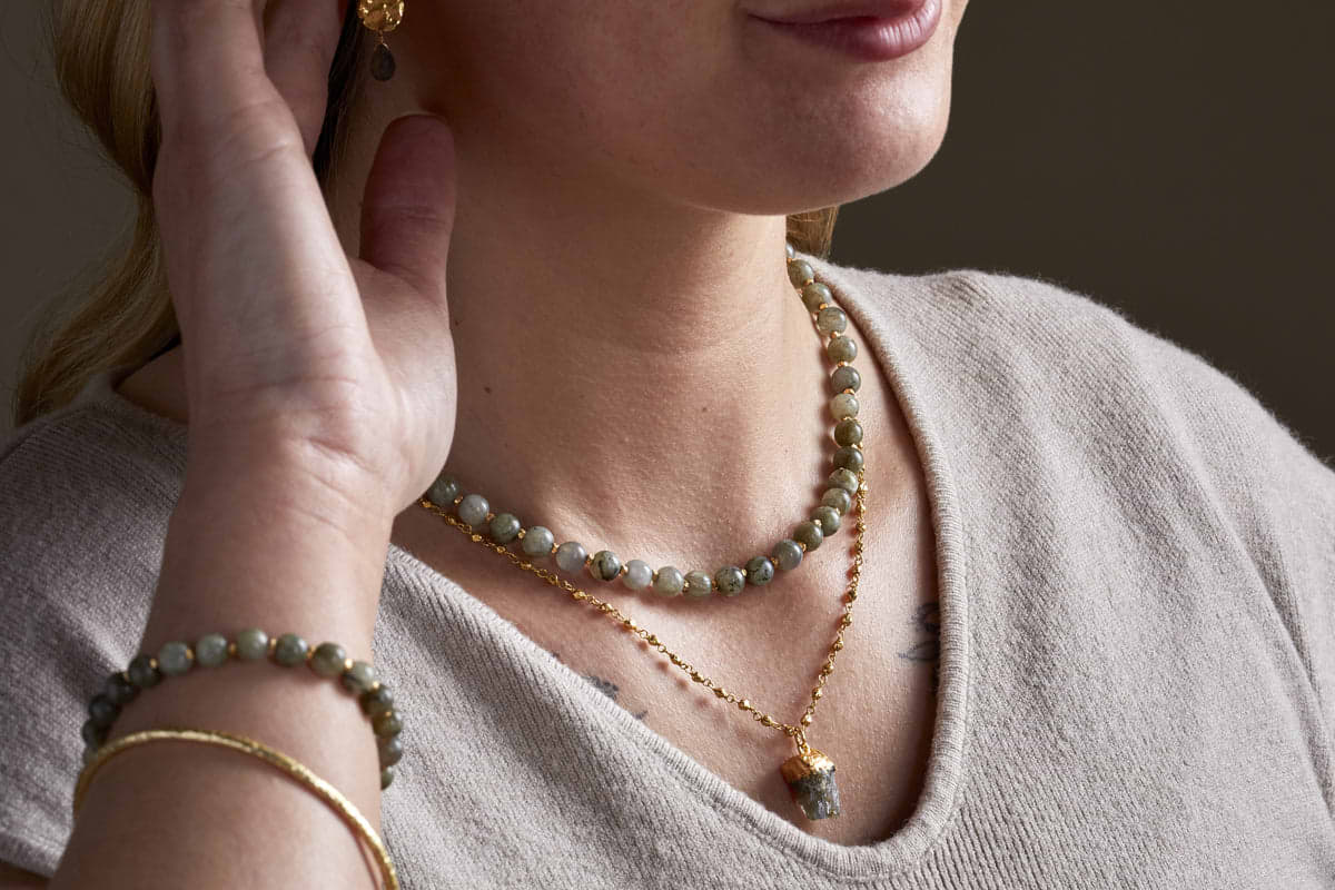 A close-up of a woman wearing a light beige top, the nkuku Hadari Labradorite Necklace in gold, a matching beaded bracelet, a gold pendant necklace, a gold bangle, and partially visible gold earrings as she touches her ear.