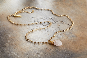 The nkuku Hadari Rose Quartz Necklace features a delicate gold chain with subtle beads and a rough teardrop-shaped rose quartz pendant, elegantly displayed on textured light brown stone—perfect for any occasion.