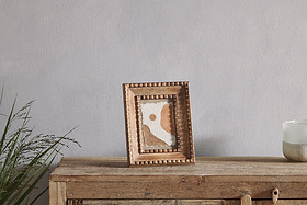 A wooden cabinet displays the nkuku Hansika Mango Wood Frame - Natural, showcasing abstract brown, white, and tan art. A green plant sits on the left and a white candle in a glass holder on the right, with a light gray wall behind.