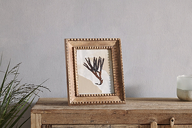 A rustic table displays an abstract painting of dark branches on beige and white, set in the Hansika Mango Wood Frame - Natural by nkuku, which features a hand-carved beaded edge. A green plant is on the left; to the right is a candle in a white glass holder.