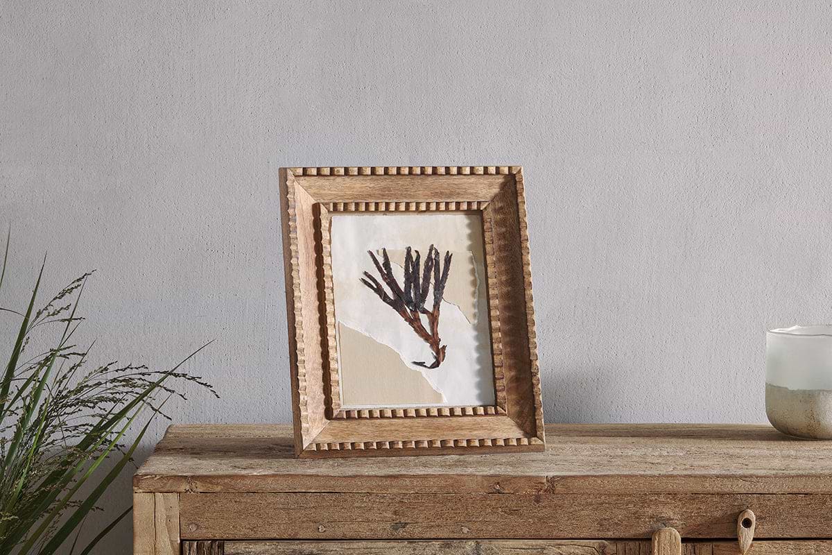 A rustic table displays an abstract painting of dark branches on beige and white, set in the Hansika Mango Wood Frame - Natural by nkuku, which features a hand-carved beaded edge. A green plant is on the left; to the right is a candle in a white glass holder.
