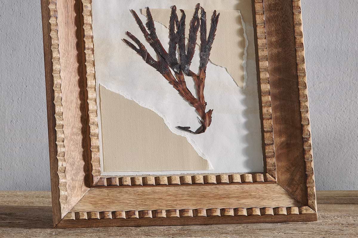 The Hansika Mango Wood Frame - Natural by nkuku features a hand-carved mango wood design with a ridged, beaded border, displaying minimalist dried plant art on layered cream and beige paper, set on a wooden surface against a light gray wall.