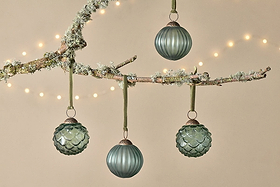 The Hara Baubles - Green (Set of 4) by nkuku, crafted from recycled glass and hung on green ribbons, adorn a frosted tree branch with moss and snow. Warm white string lights add a festive touch against a beige wall for Christmas decor.
