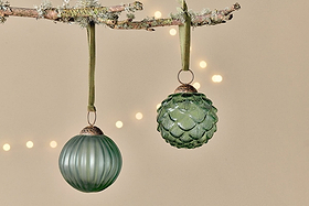 Two green Hara Baubles by nkuku hang from a lichen-covered branch on green ribbons. One bauble is ribbed while the other has a scalloped, pinecone design. Warm fairy lights glow softly in the blurred beige background.