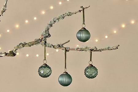 A frost-dusted branch displays three nkuku Hara Baubles - Green (Set of 4), crafted from recycled glass with unique textured patterns. Soft string lights and a beige backdrop create a festive, cozy vibe ideal for Christmas decor.