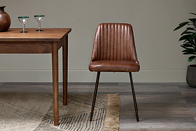 The nkuku Harsha Leather Dining Chair in chocolate brown sits beside a wooden table with two glass goblets, atop a beige fringed rug. A potted plant is on the right, set against neutral-toned walls and flooring.