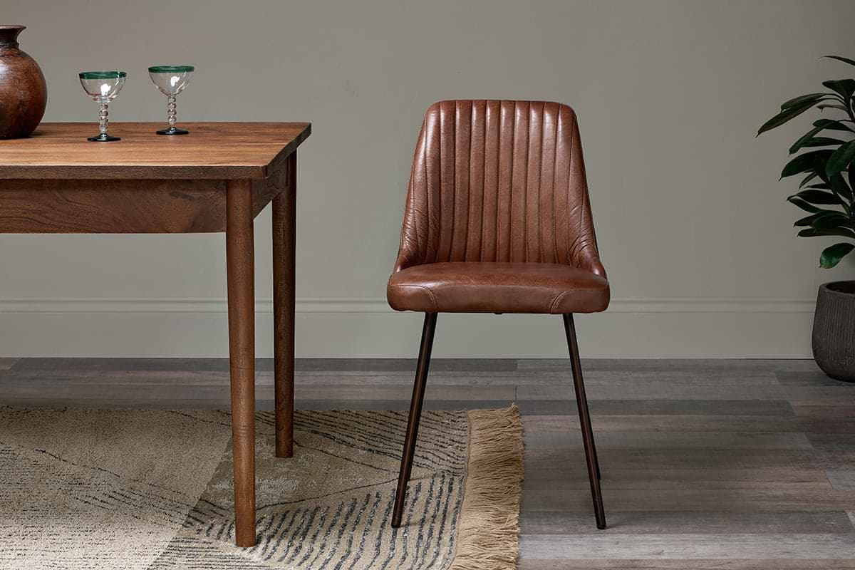 The nkuku Harsha Leather Dining Chair in chocolate brown sits beside a wooden table with two glass goblets, atop a beige fringed rug. A potted plant is on the right, set against neutral-toned walls and flooring.