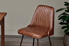 The Harsha Leather Dining Chair by nkuku in chocolate brown, with sleek dark metal legs, sits beside a wooden table. A green plant to the right brings a fresh accent to the setting.