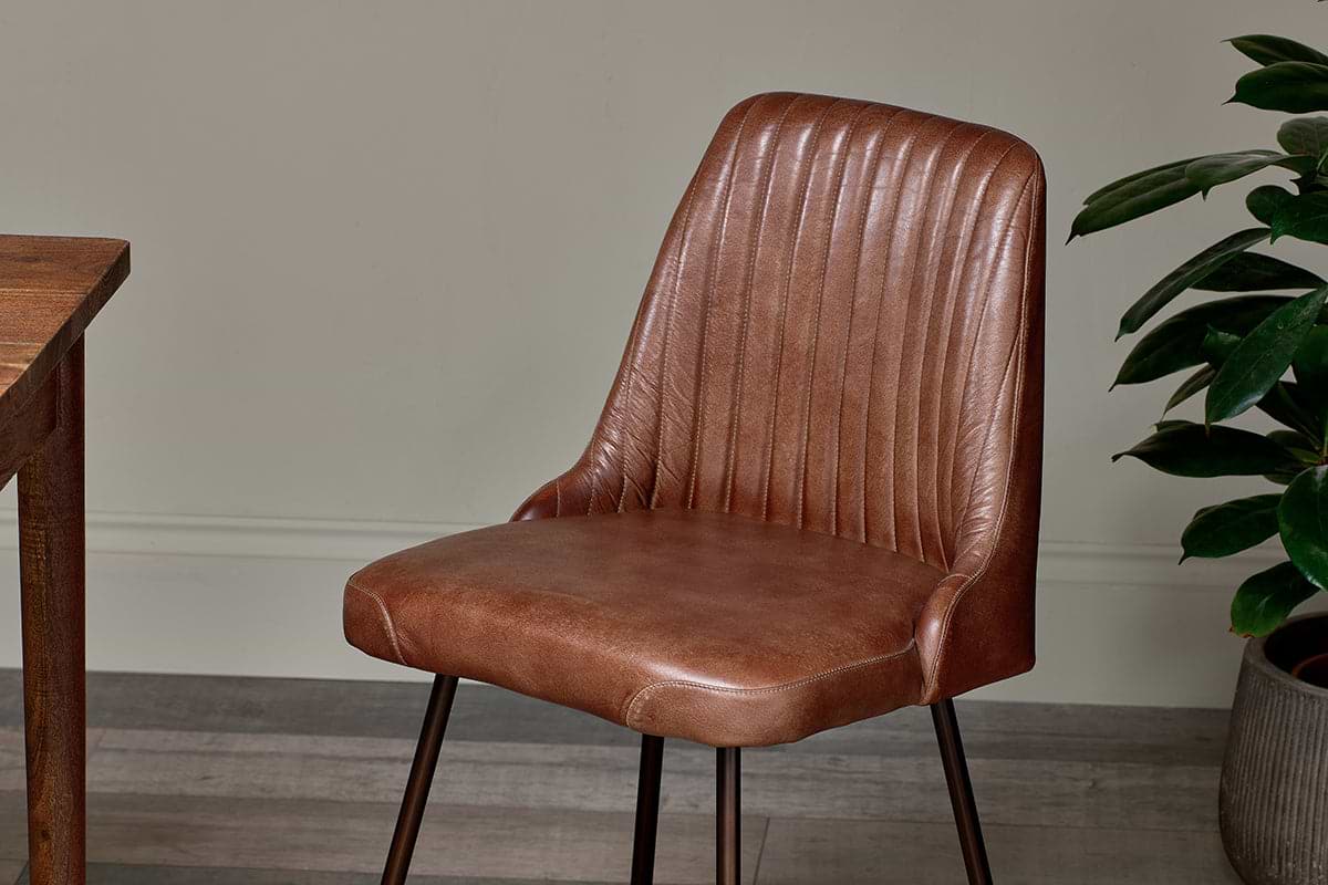 The Harsha Leather Dining Chair by nkuku in chocolate brown, with sleek dark metal legs, sits beside a wooden table. A green plant to the right brings a fresh accent to the setting.