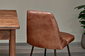 A close-up shows the back of the nkuku Harsha Leather Dining Chair in chocolate brown, with a wooden table on the left.